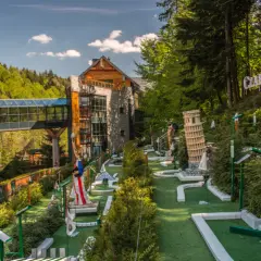 Mini golf z modelami znanych budowli, budynkiem i kładką w lesie.