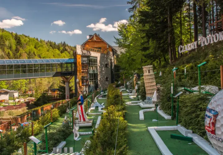 Mini golf z modelami znanych budowli, budynkiem i kładką w lesie.