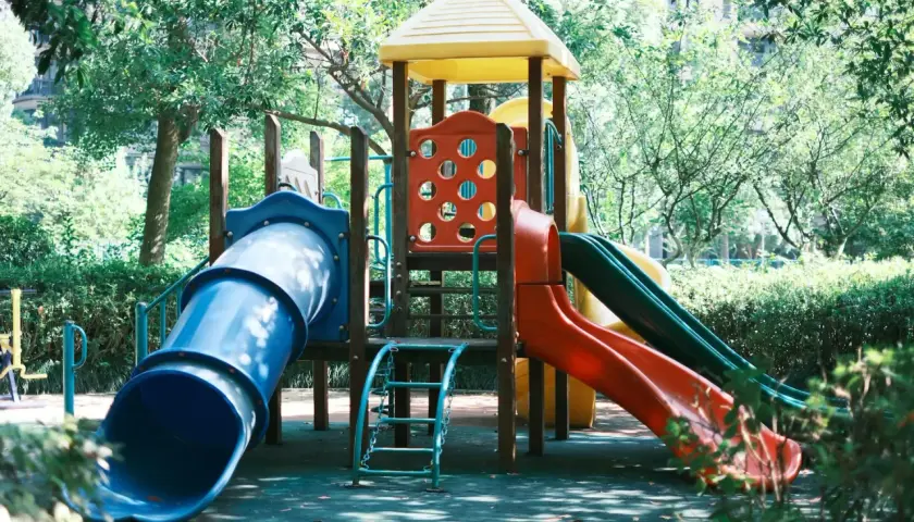 Kolorowy plac zabaw z niebieską i czerwoną zjeżdżalnią, otoczony zielenią.