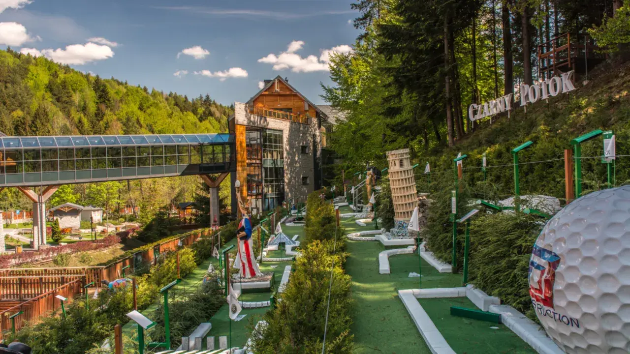 Mini golf z modelami znanych budowli, budynkiem i kładką w lesie.